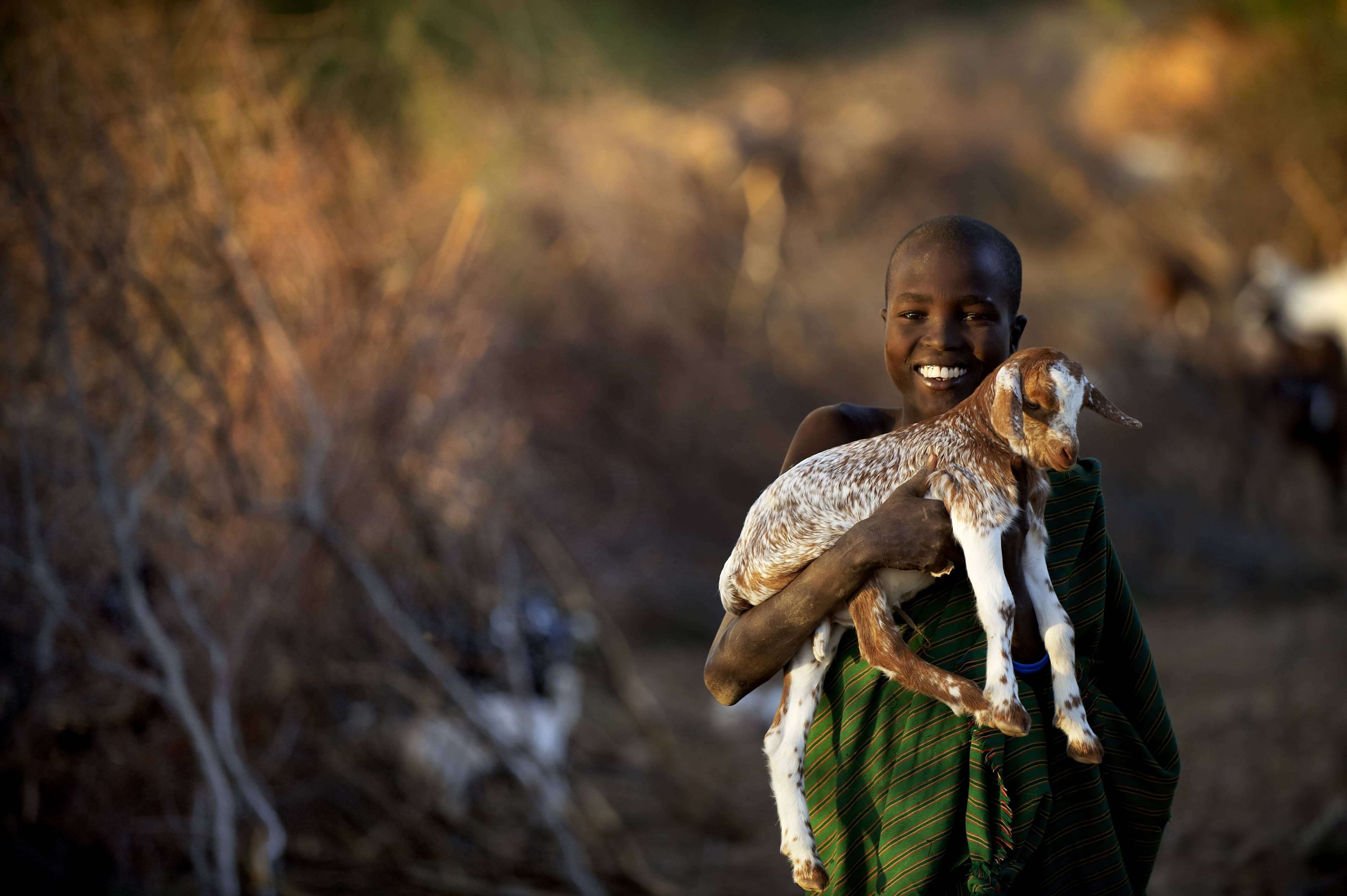 Smiling child in a green shawl holding a young goat against a natural background.
